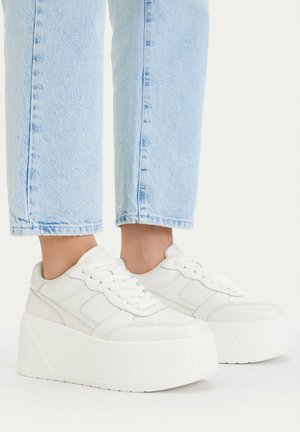 Pies con vaqueros azul claro y zapatillas blancas de plataforma con suelas gruesas y texturizadas sobre un fondo blanco.