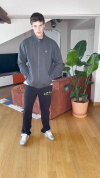 Fiatal férfi szürke cipzáras dzsekiben, fekete nadrágban és ezüstcipőben áll, a kezei a zsebében, egy nappaliban, ahol növény és narancsszínű kanapé található.