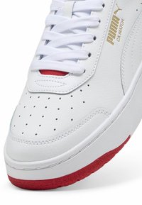 Hvide lædersneakers med perforeret tå, hvide snørebånd, røde detaljer på hælen og guld logo på siden. Glat tekstur og klassisk design.