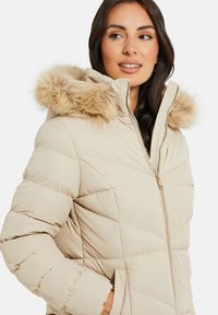 Beige Pufferjacke mit einer Kapuze aus Kunstfell, diagonalen Nähten, Reißverschluss, seitlichen Taschen und einer glatten, matten Textur.