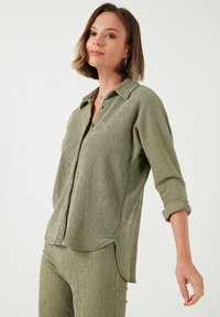 Chemise boutonnée en vert olive, texturée, à manches longues, avec un col et un ourlet arrondi, présentant des boutons visibles et une coupe décontractée.