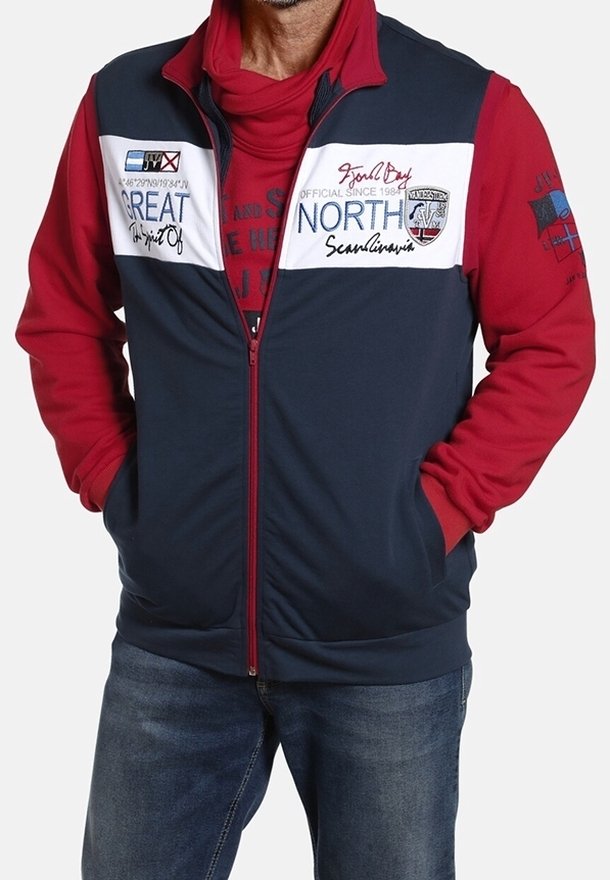 Homme portant un sweat à capuche rouge sous une veste zippée bleu marine et blanche avec texte et écussons, associé à un jean bleu, mains dans les poches.