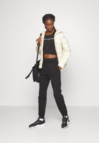 Chaqueta acolchada crema, top corto negro con el texto "Champion", pantalones deportivos negros y zapatillas negras. Materiales texturizados y colores contrastantes.