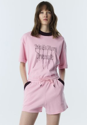 Jong persoon met schouderlang haar die een roze shirt draagt met zwarte tekst "Neem me mee waar dromen gaan" en bijpassende roze short met trekkoorden.