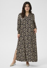 Maxikleid in Beige mit einem schwarzen geometrischen Würfelmuster. Hat lange Ärmel, einen V-Ausschnitt und seitliche Schlitze. Glatte Textur.