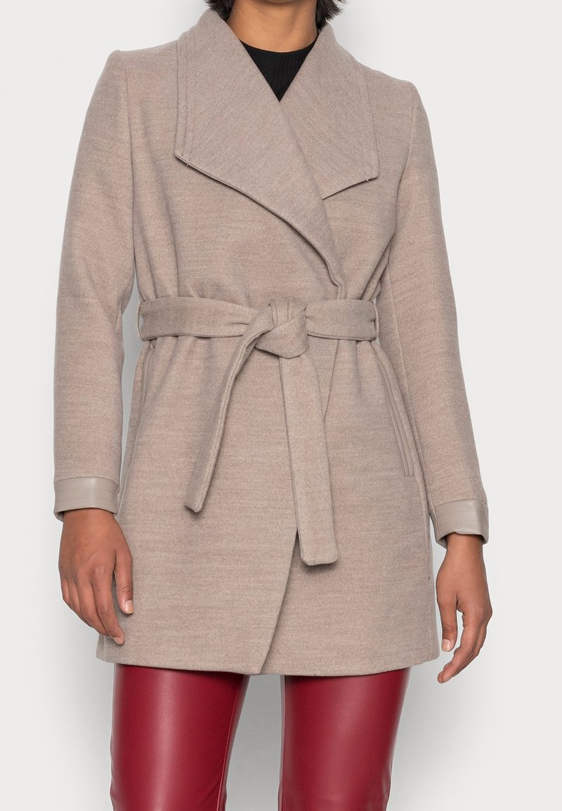 Ljust beige kappa med en omslagsdesign, bred krage och ett knytbälte. Tillverkad av mjukt tyg, med långa ärmar och kontrasterande manschetter.