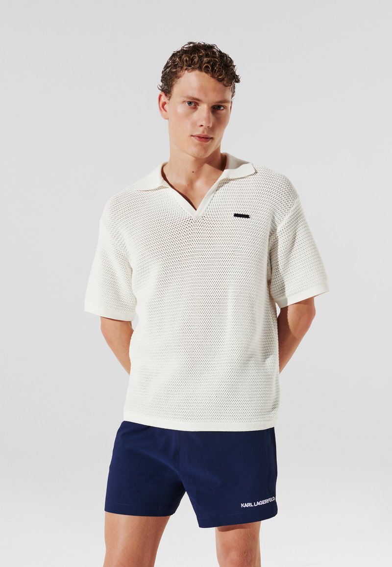 Tricou polo cu mâneci scurte, de culoare crem, cu un model texturat din plasă și decolteu în V. Are un logo negru contrastant pe piept.