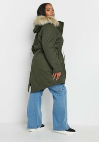 Grön parka med huva i konstgjort päls, dragsko i midjan och sidofickor, matchad med ljusblå vida jeans och svarta slip-on sneakers.