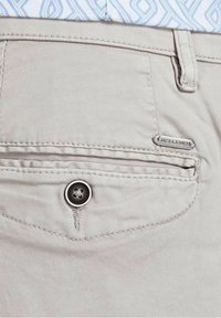 Světle béžové kalhoty se zadní kapsou se zapínáním na knoflík. Obsahují malou stříbrnou štítku s nápisem "JACK & JONES." Hladký textura látky.