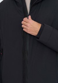 Schwarze Jacke mit Reißverschluss vorne, weicher Stofftextur und Strickbündchen. Hand nahe dem Reißverschluss zur Demonstration des Verschlusses positioniert.