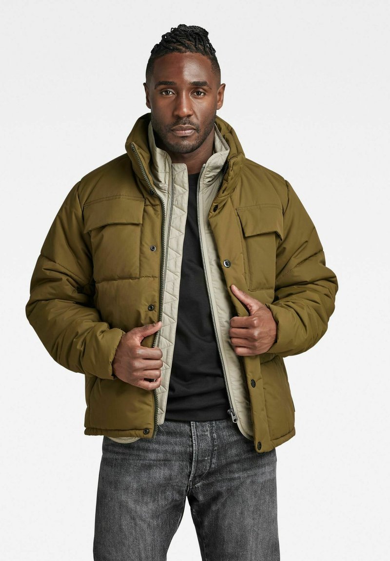 Man in gelaagd olijfgroen en beige gewatteerde jassen over een zwart shirt en grijze jeans, die de ritssluiting van de buitenste jas aanpast.