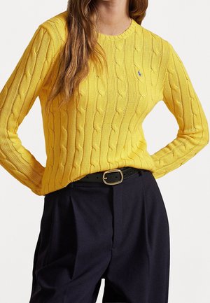 Frau trägt einen gelben, kabelgestrickten Pullover, der in eine hoch taillierte schwarze Hose mit schwarzem Gürtel gesteckt ist, steht mit den Händen in der Hüfte.