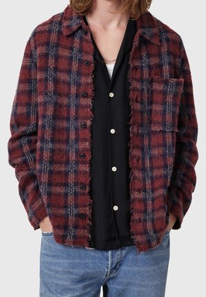 Rød og marineblå ternet jakke med knapper over en sort skjorte med knapper og lyseblå jeans, hænder i lommerne, mellem-langt hår synligt.