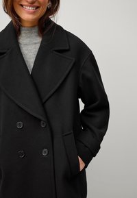 Abrigo negro de doble botonadura con solapa ancha, grandes botones y bolsillos laterales, hecho de una tela suave y con un ajuste entallado.