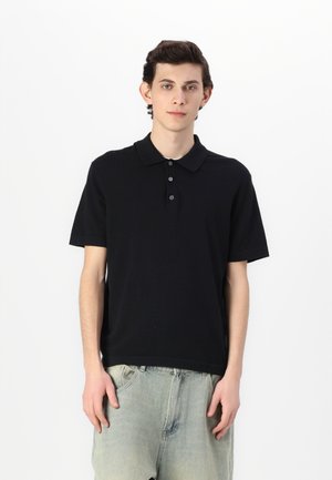 Polo noir classique avec col et patte de boutonnage à trois boutons, associé à un pantalon en denim clair, sur fond blanc.
