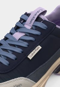 Αθλητικό παπούτσι Calvin Klein με μπλε κορδόνια, γλωττίδα σε λιλά χρώμα, ναυτικό μπλε πάνω μέρος και μπεζ σόλα, με ετικέτες της μάρκας στο πλάι και στη γλώττα.