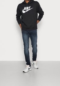 Sudadera negra de Nike con logo blanco, combinada con jeans azul oscuro y zapatillas blancas con detalles en negro. Diseñada para uso casual.