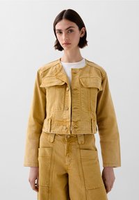 Giacca in denim gialla tagliata, con due tasche anteriori a pattina, chiusura a bottone e una finitura leggermente texturizzata. Indossata sopra un top bianco.