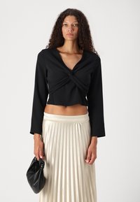 Svart langermet topp med en vridd front, kombinert med en plisserte kremfarge skjørt og holder en liten svart håndveske. Glatt tekstur av stoffet.