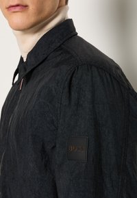 Homme portant une veste noire texturée avec un petit patch logo BOSS sur le haut du bras, associée à un pull col roulé beige en dessous.