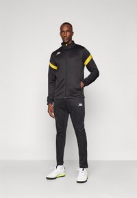 Black track jacket with yellow accents, zip closure, and logo; paired with black pants. Model wears white shoes with green highlights.