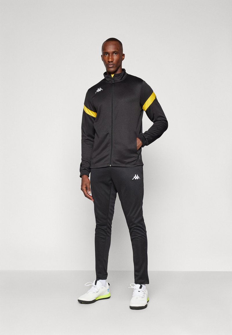 Black track jacket with yellow accents, zip closure, and logo; paired with black pants. Model wears white shoes with green highlights.