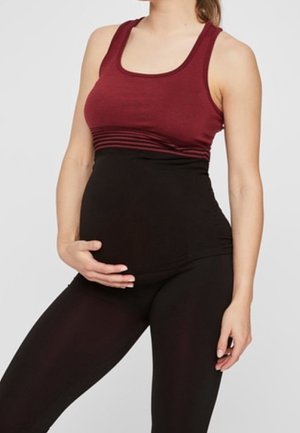 Femme enceinte portant un haut sans manches bordeaux et des leggings de maternité noirs, tenant son ventre avec une main.