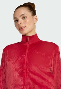 Crvena zip-jakna izrađena od mekog baršuna. Ima visoki ovratnik i utisnuti detalj s logom. Glatka tekstura s laganim sjajem.