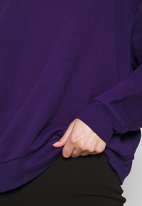 Sudadera morada con mangas largas, corte relajado, puños acanalados y una textura suave, sostenida por una mano en el dobladillo.