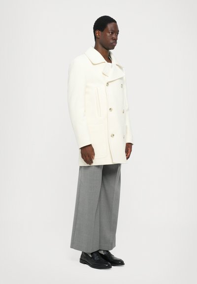Cappotto in lana bianco con design doppiopetto, ampio colletto e tasche laterali. Abbinato a pantaloni grigi a gamba larga e scarpe nere.