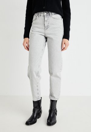 Personne portant un jean taille haute gris clair, un pull noir à manches longues et des bottines en cuir noires, debout devant un fond uni.