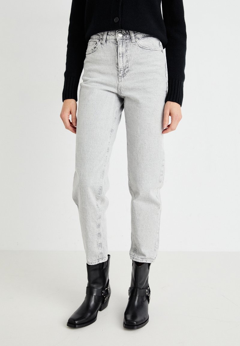 Personne portant un jean taille haute gris clair, un pull noir à manches longues et des bottines en cuir noires, debout devant un fond uni.