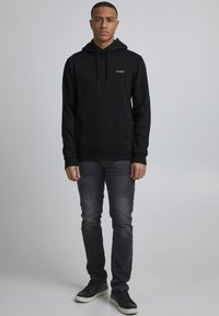 Černá mikina s kapucí a klokaní kapsou, texturovaná tkanina a decentní logo na prsou, v kombinaci s tmavě šedými slim-fit džíny a černými teniskami.