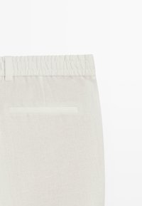 Gros plan sur un pantalon en lin beige doté d'une taille élastique et d'une unique poche arrière horizontale avec une ouverture passepoilée.