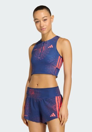 Kobieta w niebiesko-różowym sportowym crop topie w kwiatowy wzór i pasujących do niego szortach, stojąca w luźnej pozie na jednolitym tle.