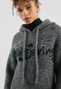 Woman with curly hair wears a gray knitted hoodie with black text "Breaking free" and "Daytime" over a black turtleneck.