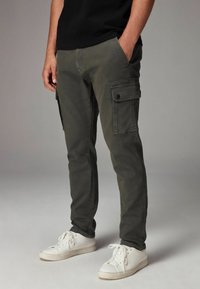 Olivgrüne Cargo-Hosen mit seitlichen Taschen, schmaler Schnitt und zulaufenden Beinen. Getragen mit weißen Sneakern, die über eine glatte, robuste Textur verfügen.
