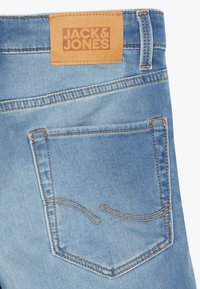 Jeans de mezclilla azul claro con un parche de cuero marrón claro etiquetado "JACK & JONES". Presenta un bolsillo trasero con costuras curvas destacadas.