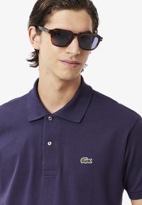 Ung man med mörkt hår som bär solglasögon och en lila polo-tröja med en liten grön krokodilmärke, ler självsäkert mot kameran.