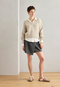 Μπεζ πλεχτό πουλόβερ με χαλαρή εφαρμογή, τοποθετημένο πάνω από μια λευκή πουκαμίσα με γιακά. Σκούρα γκρι φούστα με ρίγες και ανοιχτό ροζ σαμπάνιας σάνδαλα με λουλούδια.