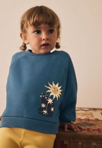 Blå bomullströja med en sol- och kattgrafik i gult och svart, bärs med gula byxor; har ribbad halsringning och muddar.