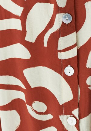 Blouse en coton avec un fond rouge rouille orné de grands motifs abstraits blancs, et des boutons en perle le long de l'avant.