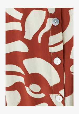 Blouse en coton avec un fond rouge rouille orné de grands motifs abstraits blancs, et des boutons en perle le long de l'avant.