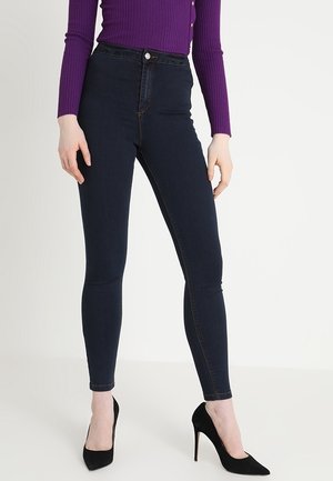 Personne portant un jean skinny bleu foncé, un haut à manches longues violet côtelé, et des chaussures à talons hauts noires, debout devant un fond blanc.