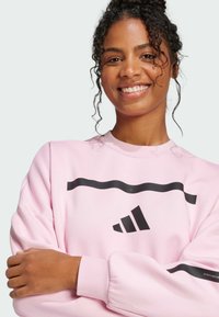 Light pink sweatshirt with black striped detailing and logo. Features crew neck, long sleeves, and textured fabric. Simplistic design.