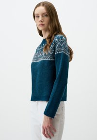 Turquoise trui met een patroon op de schouderpartij in wit en lichtblauw. Zachte stof, ronde hals en geribde manchetten, gedragen met witte broek.