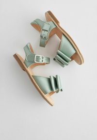 Sandali in raso verde con design a strap e fibbia, caratterizzati da un grande dettaglio a fiocco su un lato e una suola in gomma color beige chiaro.