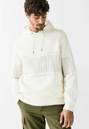 Cream hooded sweatshirt featuring embossed text, drawstring closure, and ribbed cuffs. Paired with olive green cargo pants.