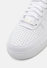 White leather sneaker featuring a woven texture on the toe box, flat white laces, and a small metallic Nike logo on the tongue.