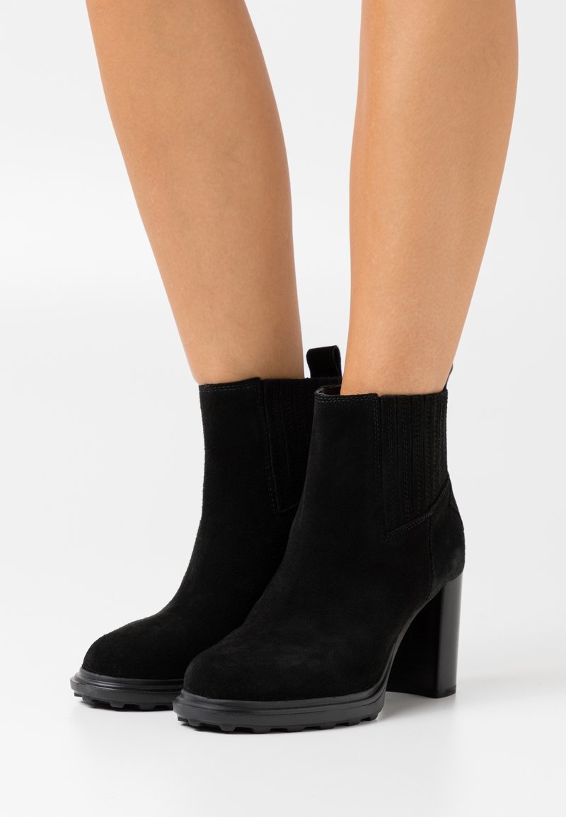 Bottes chevilles en daim noir à talons épais portées sur des jambes nues sur fond blanc.